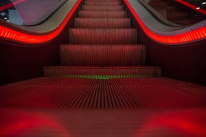An escalator, marked by numerous past accidents, with glowing red lights along the edges and steps, creating an atmospheric and moody ambiance in a dimly lit environment.