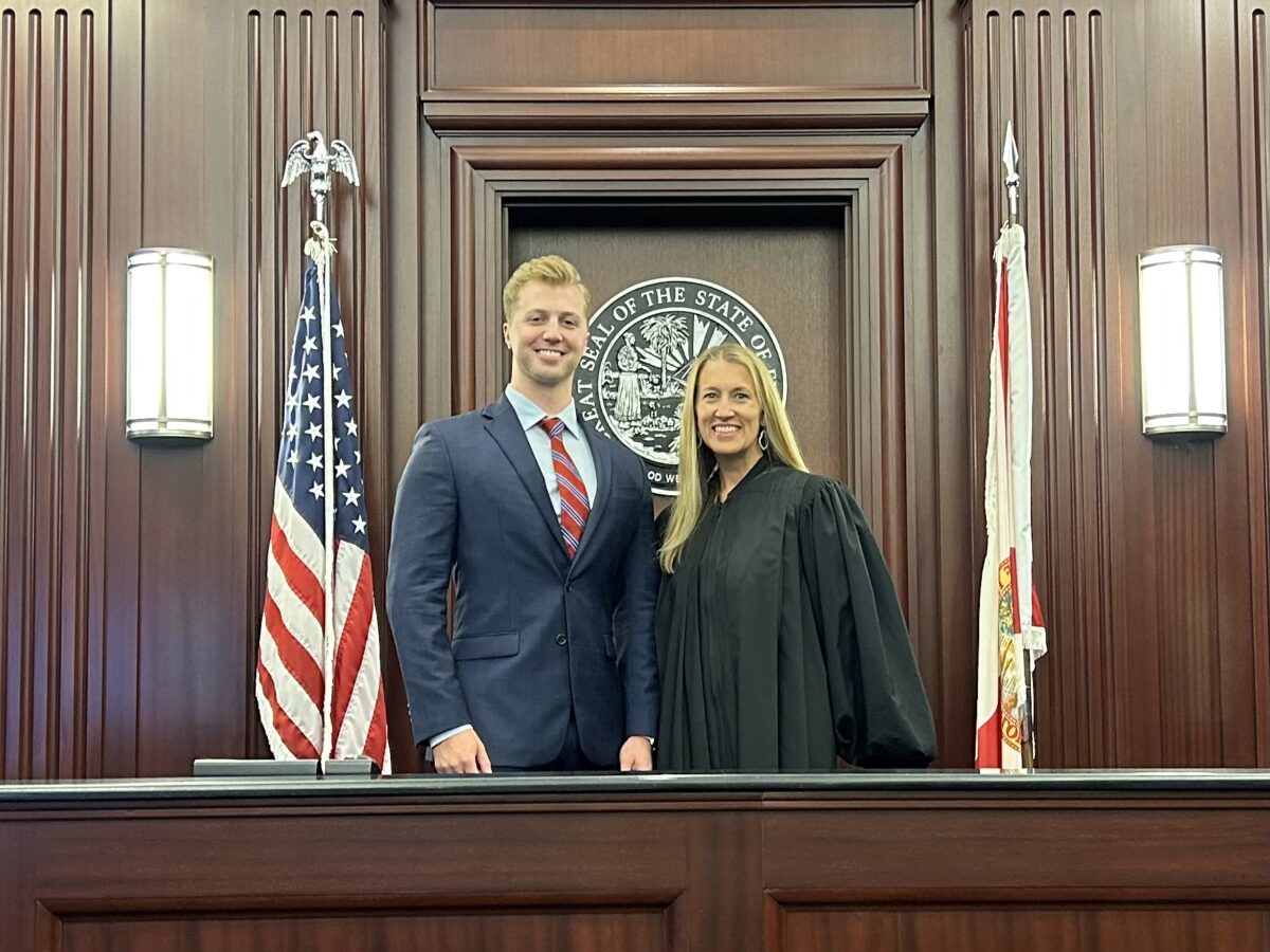 A man in a suit and a woman in a judge's robe stand side by side in the courtroom of Pairitz Ansbacher Law. Behind them, the American flag, a state flag, and a state seal adorn the wooden wall. Smiling directly at the camera, they embody professionalism and justice.